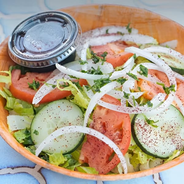 A fresh Mediterranean salad with lettuce, tomatoes, cucumbers, onions, and a side of dressing.