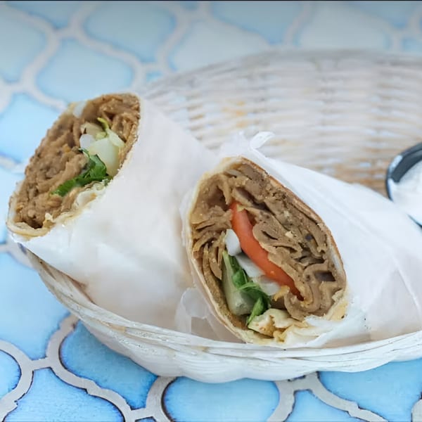 Two Bluzone Cafe Mediterranean wraps, one with tomato, in a white basket on a blue patterned table.