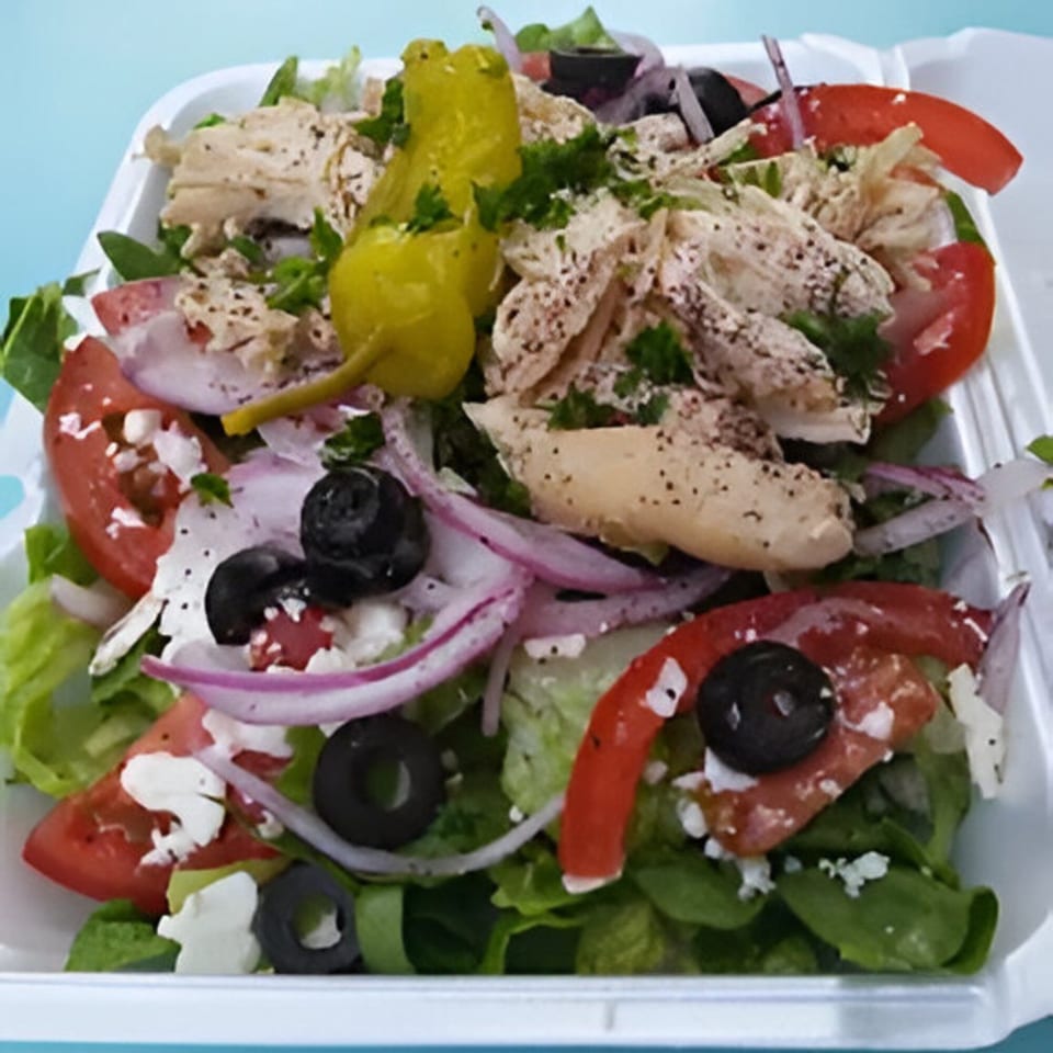A Mediterranean salad with chicken, tomatoes, olives, feta, red onion, and pepperoncini in a white container.