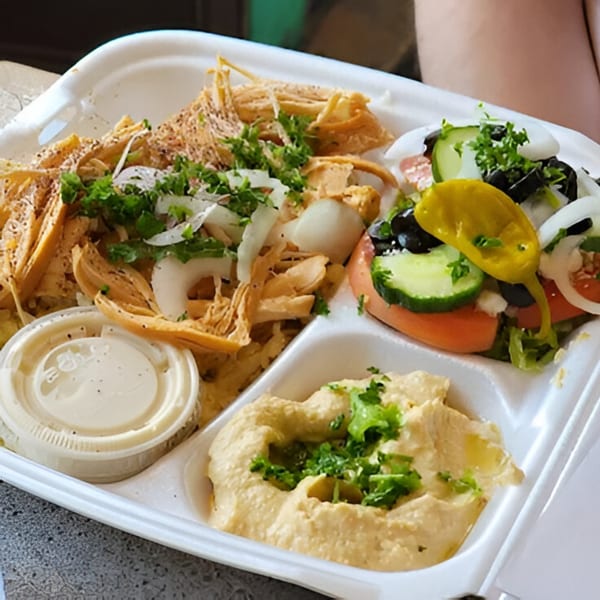 Mediterranean platter with shredded chicken, hummus, a side salad, and dressing in a white to-go container.